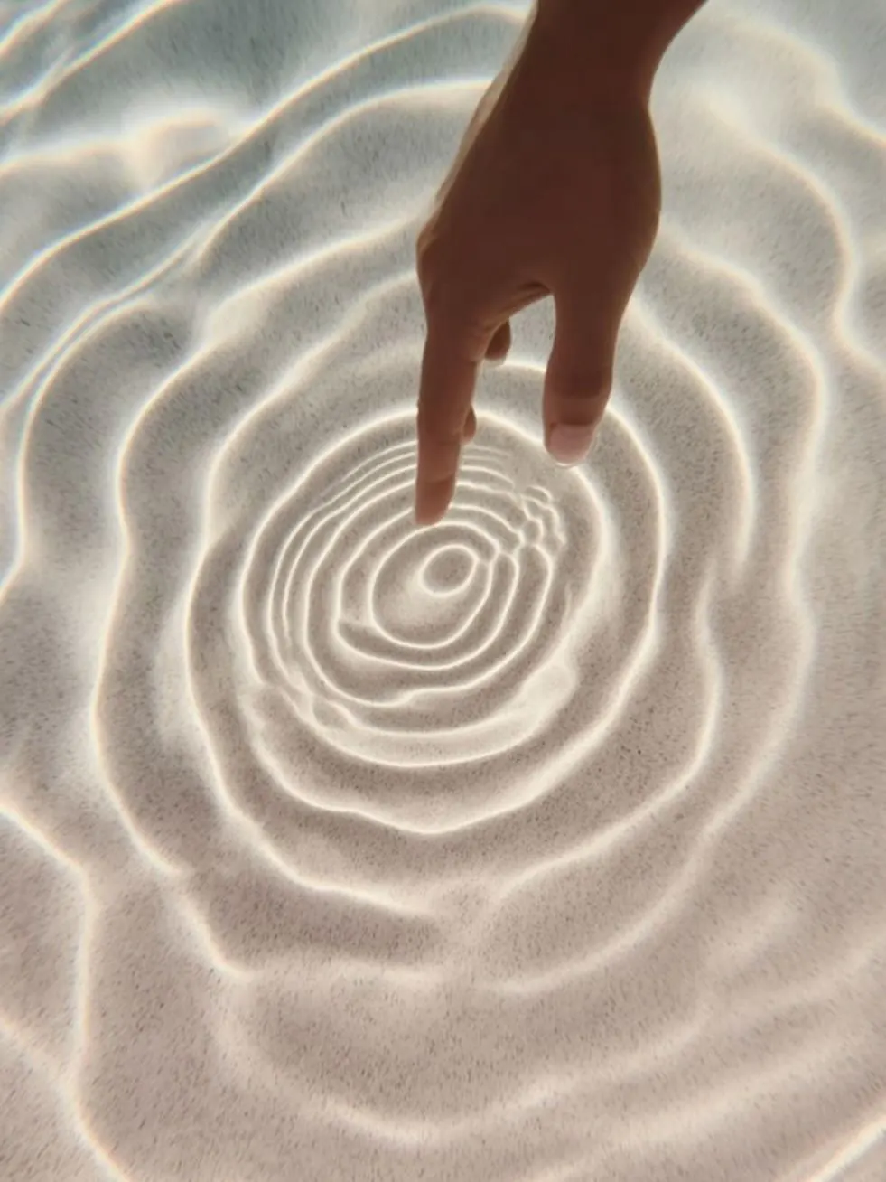 A hand touching clear water, sending ripples across the sand below